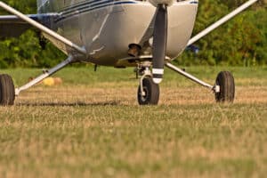 Front view of a small single-engine airplane with propeller, landing gear down, on a grassy airfield with trees in the background.